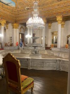 A grand hall inside Dolmabahçe Palace in Turkey, showcasing a magnificent crystal chandelier, an ornately decorated ceiling with gold details, a central marble fountain, and luxurious furniture reflecting Ottoman elegance