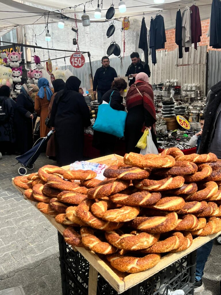 A stack of sesame-covered simit bread displayed on a wooden tray at Fatih Çarşamba Pazarı, with shoppers browsing kitchenware in the background.